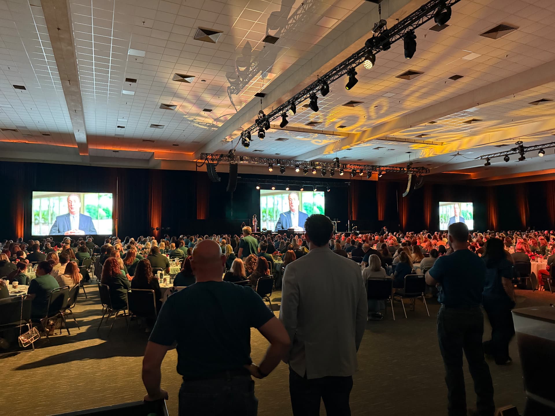 Black stage drape backdrop behind a general session with three projection screens and truss lighting