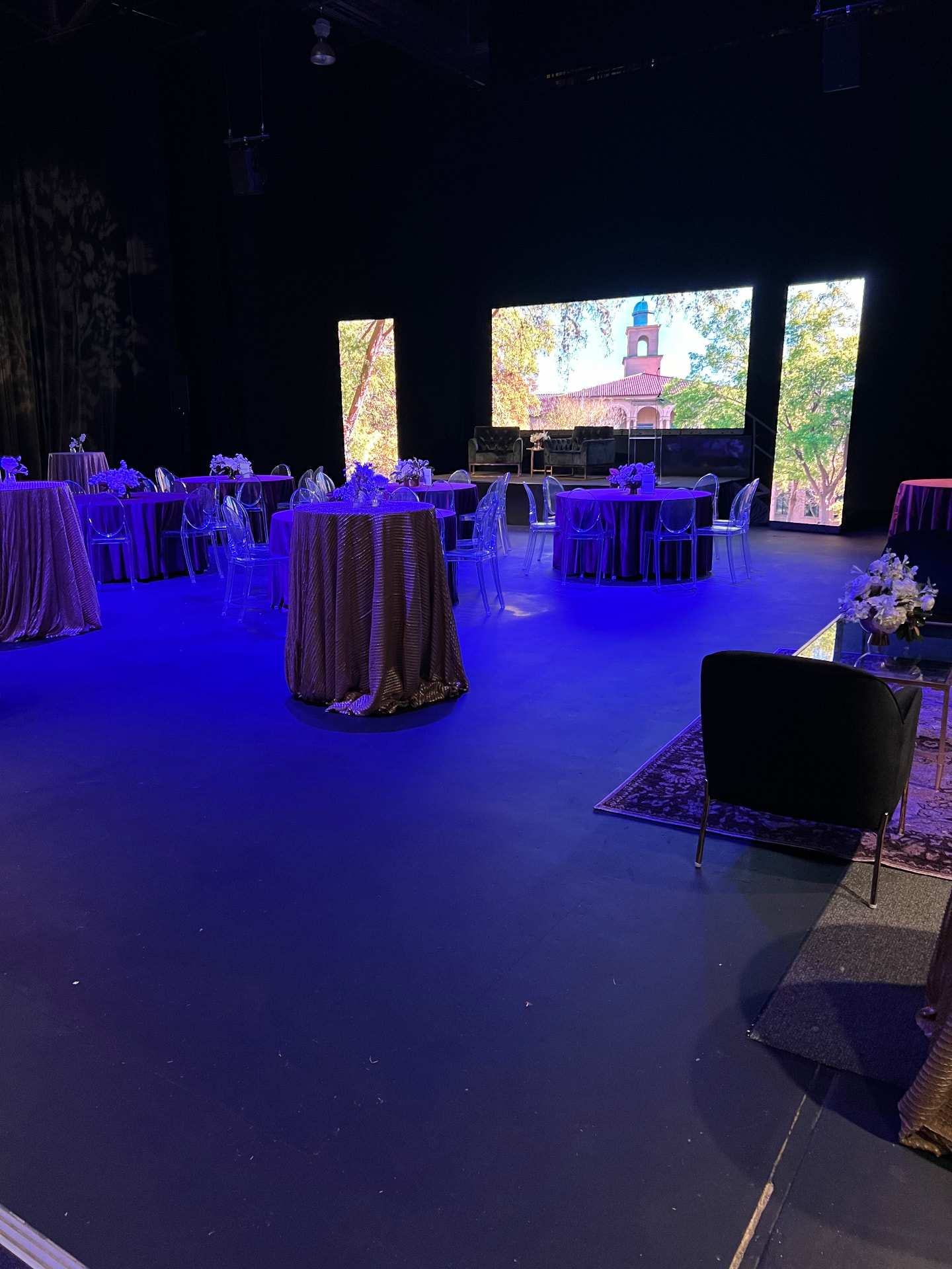 Indoor gala lit in blue with round tables and clear ghost chairs facing three large LED video walls